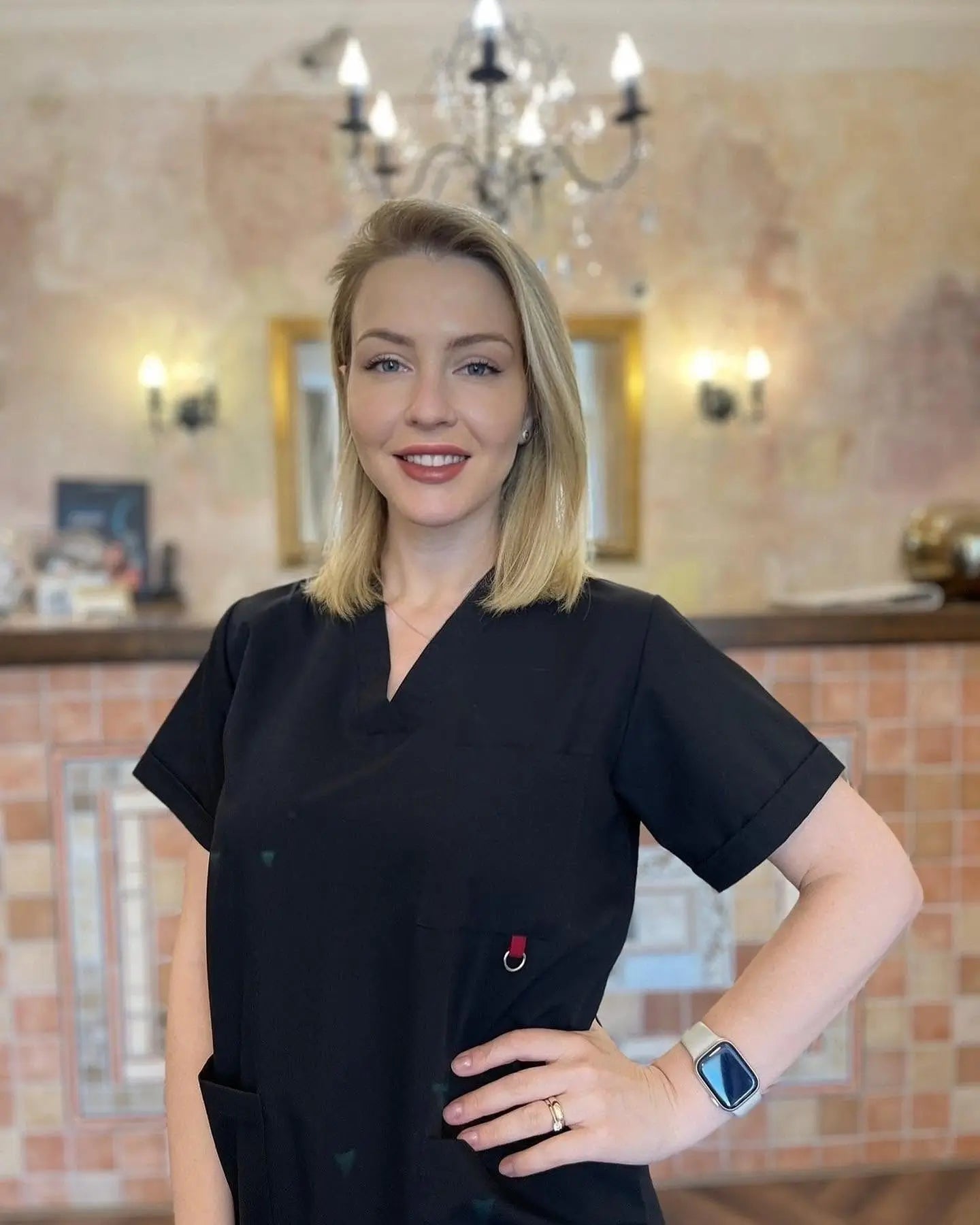 Woman in black scrub top standing in a room with a chandelier and wall lights.