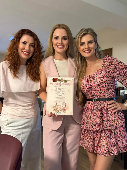 Three women standing together indoors, holding a printed certificate or award. The woman in the center wears a light pink blazer over a white top, the woman on the left wears a white top and white pants, and the woman on the right wears a pink floral dress with black and gray abstract patterning.